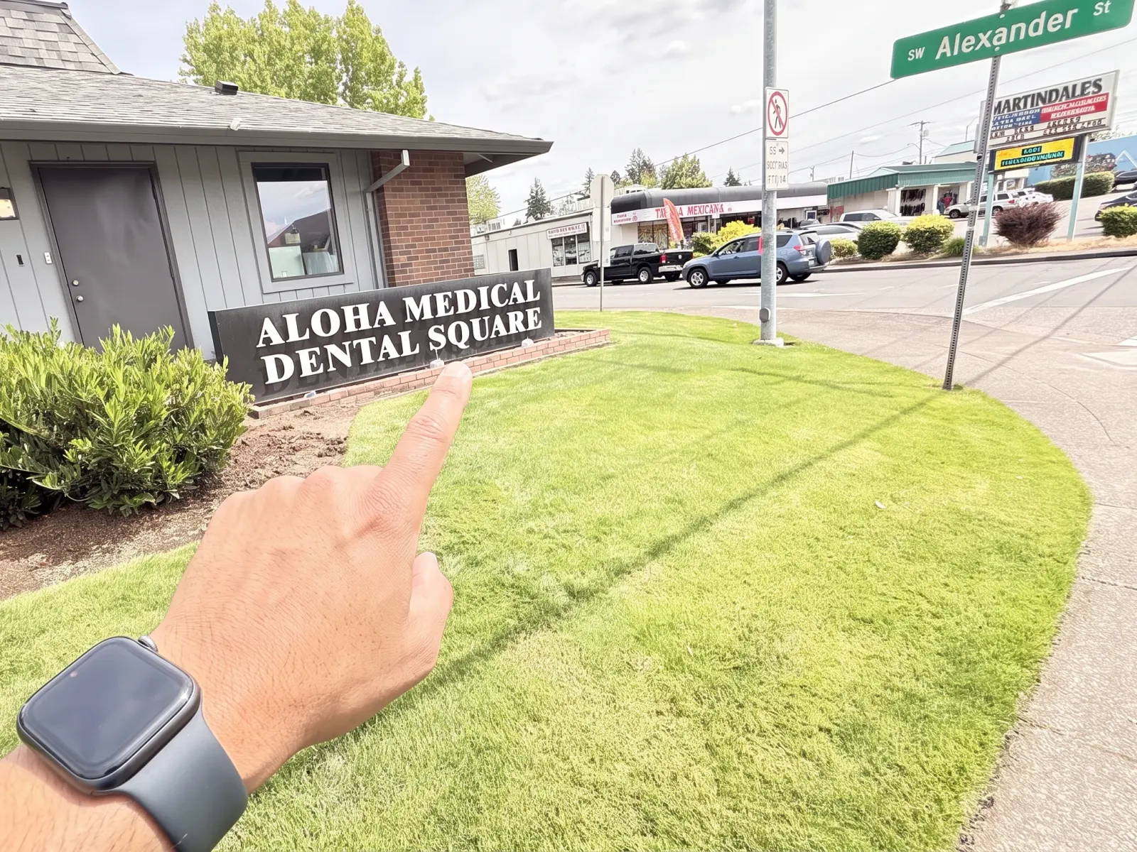 A hand points to the Aloha Medical Dental Square sign outside the Aloha Dental Specialty Center building at SW Alexander St in Beaverton, Oregon